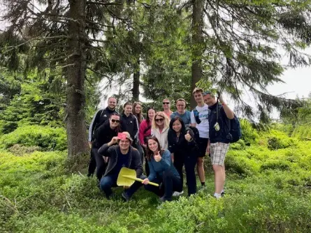 Skupina lidí stojících a posedávajících v lese mezi stromy a zelení, připraveni na venkovní aktivitu či výlet v rámci teambuildingu..