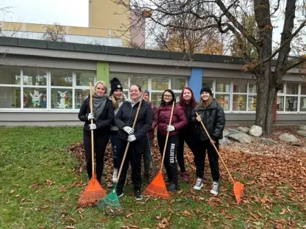 Skupina lidí venku u školní budovy, připravující se na hrabání spadaného listí, každý drží hrábě, jesení atmosféra v rámci dobrovolnického dne.