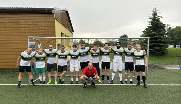 Tým jedenácti mužů ve fotbalových dresech s nápisem „MND“ stojí a klečí před fotbalovou brankou na zeleném hřišti během Fotbalového turnaje KKCG.