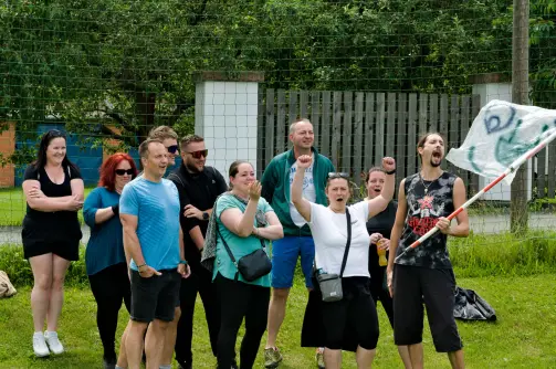 Skupina lidí venku na trávníku, někteří s nadšeným gestem, jeden drží vlajku, v pozadí plot a stromy, atmosféra teambuildingu..
