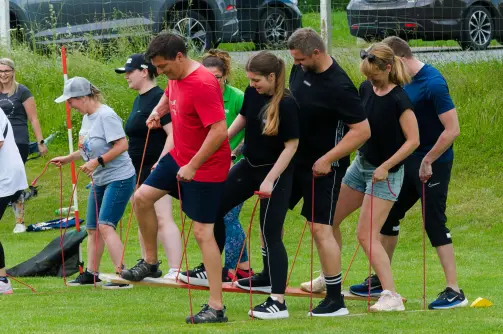 Skupina lidí na venkovní týmové aktivitě na trávníku, koordinovaně kráčející po dřevěných prknech pomocí lan, během sportovního nebo teambuildingového dne.